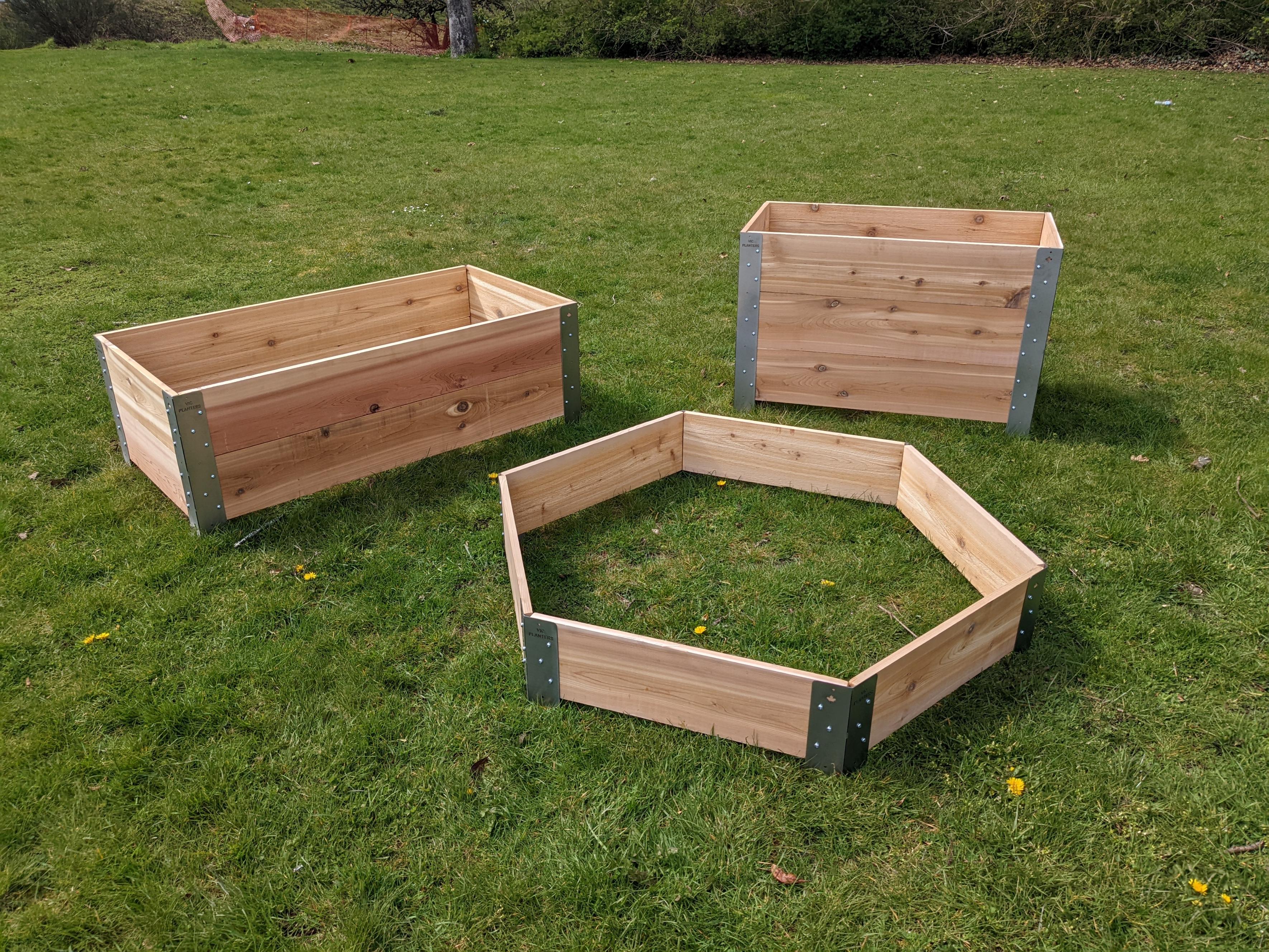 Three cedar wood planter boxes on a grass yard. In the back right is a tall rectangular box about two feet tall by three feet wide. In the back left is a short box about one foot tall and three feet wide. In front is a hexagon-shaped planter box. 